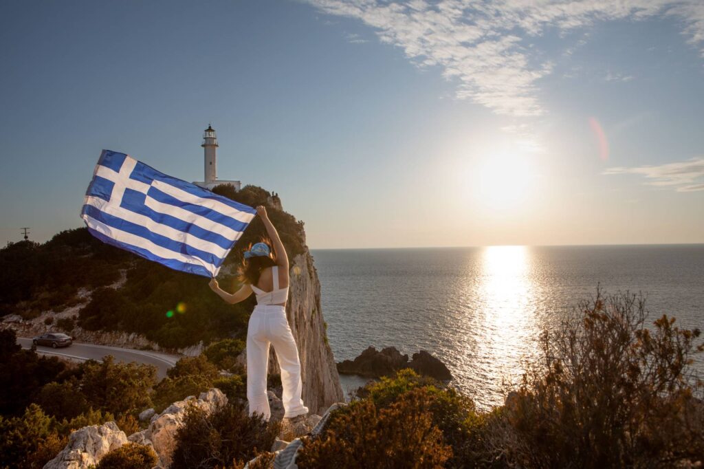 woman with greece flag looking at sunset above the sea Lefkada island lighthouse on background