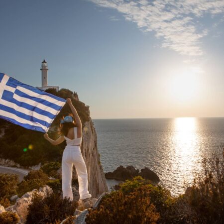 woman with greece flag looking at sunset above the sea Lefkada island lighthouse on background